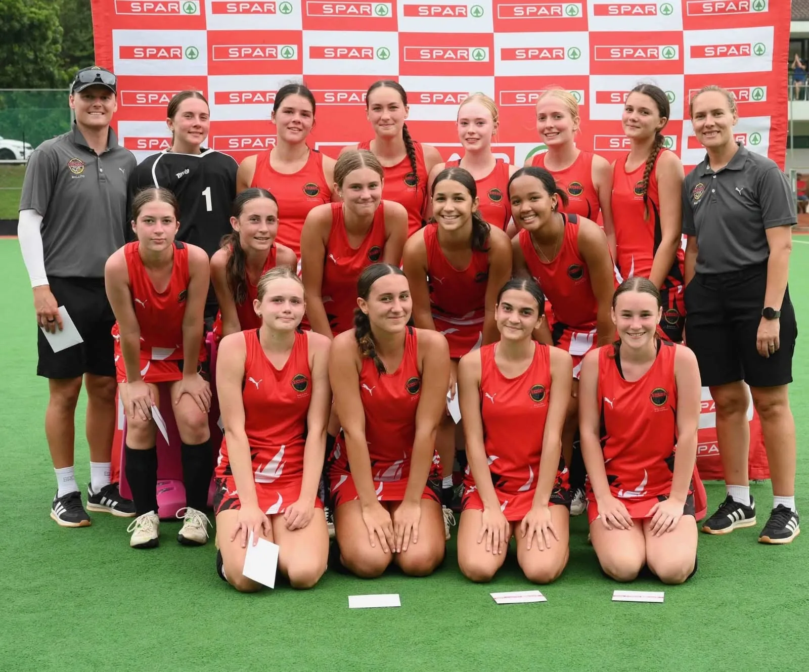 A junior girls' hockey team in red kit posing together at a tournament.